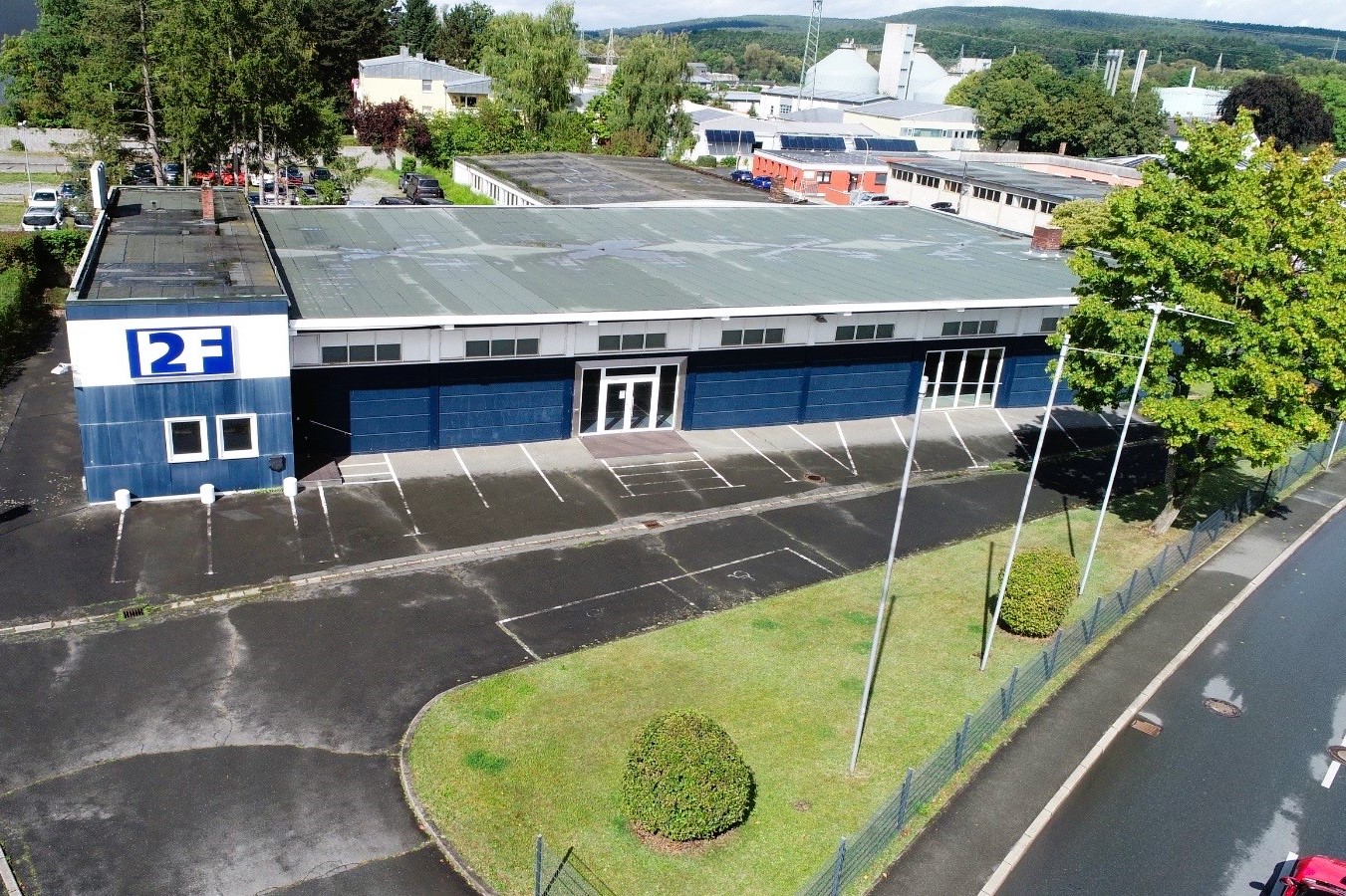 Luftbild/Schrägaufnahmen eines 1-stöckigen Hallengebäudes (dunkelblau mit weiß) mit Parkflächen und kleiner Grünfläche im Vordergund